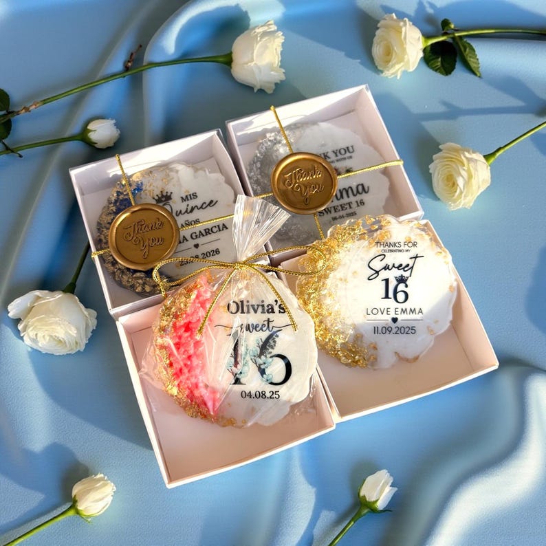 May include: Four white gift boxes with decorative cookies, gold accents, and text. The cookies have gold edges and are decorated with text such as "Sweet 16" and "Thank You". White roses are scattered around the boxes on a blue surface.