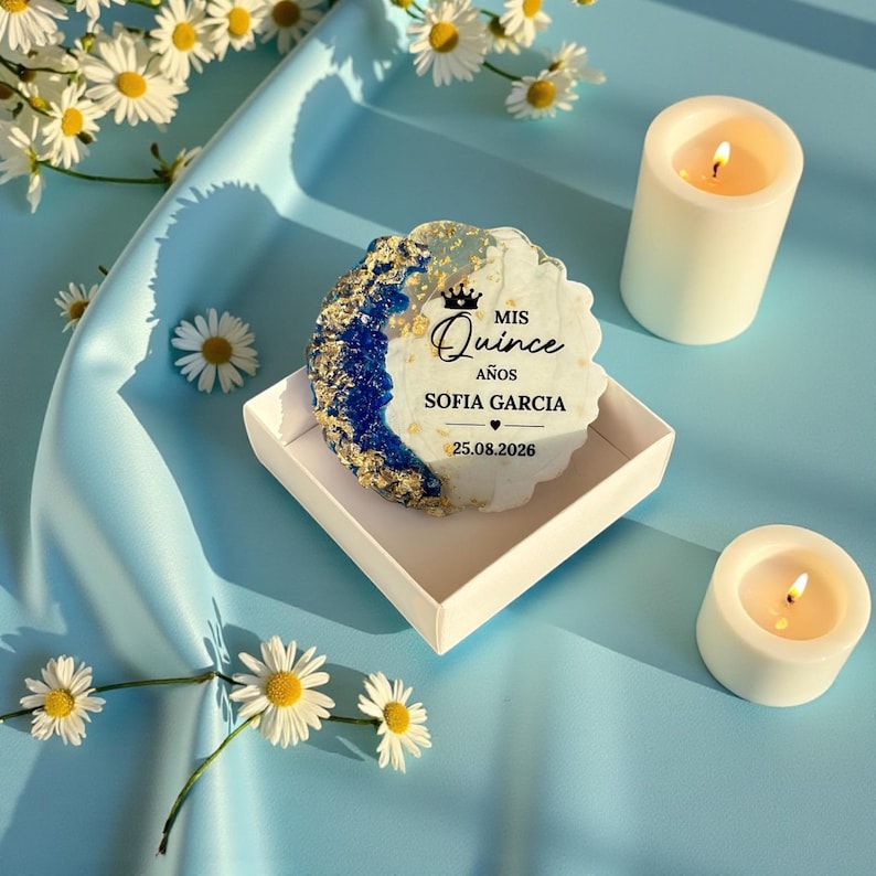 May include: A decorative item with the text "MIS Quince A&ntilde;os SOFIA GARCIA 25.08.2026" in a white box, with a blue and gold design. Two white candles are lit, and daisies are scattered around the scene.