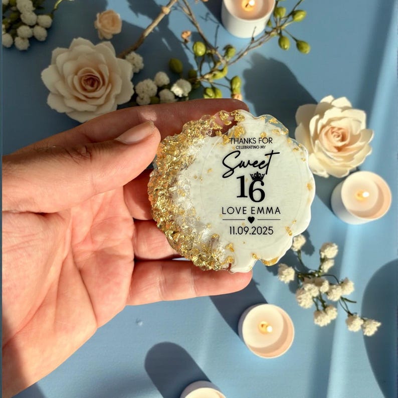 May include: A Sweet 16 keepsake with gold leaf accents and the text "Sweet 16 Love Emma 11.09.2025". The coaster is white with a gold-colored edge. The background includes white roses and candles.
