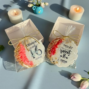 May include: Two decorated cookies in clear cellophane, tied with gold string, inside white boxes. One cookie has "Olivia's Sweet 16" text and a butterfly graphic. The other says "Sweet 16 Emma". Candles and flowers are in the background.