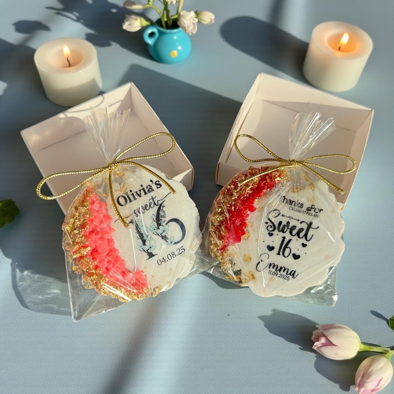 May include: Two decorated cookies in clear cellophane, tied with gold string, inside white boxes. One cookie has "Olivia's Sweet 16" text and a butterfly graphic. The other says "Sweet 16 Emma". Candles and flowers are in the background.