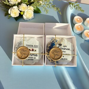 May include: Two decorative coasters in white boxes. One coaster has a blue and white design, the other has a blue and gold design. Both have a gold wax seal that says "Thank You". Text on the coasters reads "Thank You for celebrating with us" and "Sweet 16".