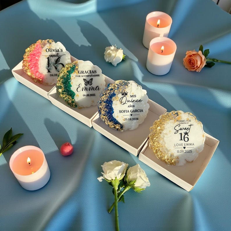 May include: A collection of decorative candles with text, including "Olivia's sweet 15", "Gracias por acompa&ntilde;arme Emma", "Mis Quince Sofia Garcia", and "Sweet 16 Love Emma". The candles are white with colorful accents and sit in individual trays. Several lit candles and flowers are also present.