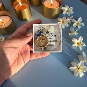 May include: A white box containing a decorative favor with blue and gold accents, a gold seal that says "Thank You", and the text "Mis Quince". The favor is tied with gold string. Several lit gold candles and white flowers are in the background.
