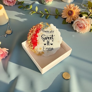 May include: A decorative coaster with a white, red, and gold design, featuring the text "Thanks For Celebrating My Sweet 16 Emma 11.09.2025". The coaster is in a white box, with flowers in the background.