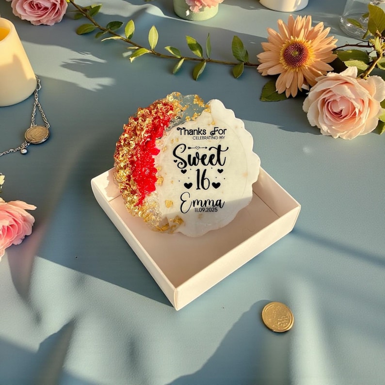 May include: A decorative coaster with a white, red, and gold design, featuring the text "Thanks For Celebrating My Sweet 16 Emma 11.09.2025". The coaster is in a white box, with flowers in the background.