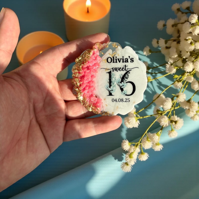 May include: A decorative coaster with a floral design and the text "Olivia's sweet 16 04.08.25". The coaster has a pink and gold border. The coaster is held in a hand, with candles and flowers in the background.