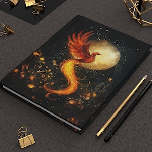 May include: A hardback journal featuring a vibrant phoenix design. The cover showcases a fiery orange and red phoenix against a dark background with a full moon and floral accents. Gold and black pencils and gold binder clips are also visible.