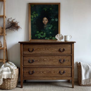May include: A dark wood dresser with three drawers and antique-style hardware. The dresser is in front of a white wall with a framed painting of a woman with dark hair in a green leafy setting above it.