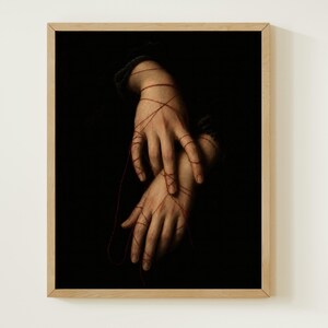 May include: A framed wall art print featuring two hands positioned against a solid black background. Thin, vibrant red threads are wrapped intricately around the fingers and wrists, creating a web-like pattern that connects the two hands together.