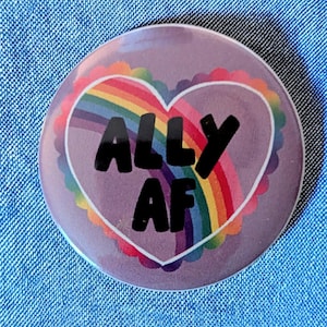 May include: A round, purple button with the text "ALLY AF" in black, inside a white heart with a rainbow and scalloped border. The button is set against a blue denim background.