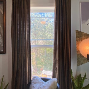 Pode incluir: Um travesseiro branco de pele sintética está em uma cadeira de couro preto na frente de uma janela com cortinas marrons. A janela dá para uma área arborizada.