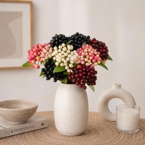 May include: A decorative arrangement of artificial berry clusters in a white ceramic vase. The berries feature a multi-coloured palette including shades of black, cream, pink, and deep burgundy, accented by small green leaves on thin stems.
