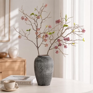May include: A dark gray, textured, cylindrical vase holds several thin, branching stems adorned with small, clustered pink flower buds and sparse, light green leaves. The vase has a weathered, cross-hatched finish and a narrow, rounded opening.