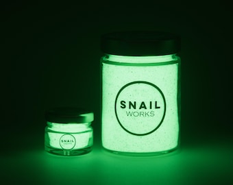 May include: Two clear glass jars of varying sizes filled with a granular substance that emits a bright green glow in the dark. Each jar features a black lid and a circular logo on the front containing the text SNAIL WORKS in black lettering.