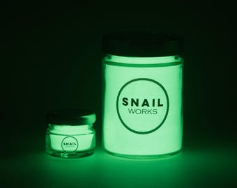 May include: Two glass jars with black lids that glow with a bright green light in the dark. Each jar features a circular logo on the front with the text SNAIL WORKS printed in black. The jars are cylindrical and contain a glowing substance.