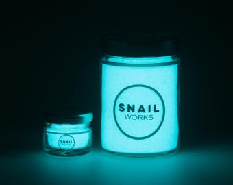 May include: Two clear glass jars filled with a glowing cyan powder. The larger jar on the right and the smaller jar on the left both feature a black circular logo with the text SNAIL WORKS printed in the center of the glowing substance.