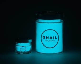 May include: Two clear glass jars with black lids containing a glowing cyan powder. The larger jar features a circular logo with the text SNAIL WORKS, while the smaller jar has a matching circular logo. Both jars emit a bright cyan light.