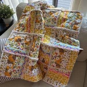 Puede incluir: Un edredón hecho a mano con un estampado floral y de mariposas. El edredón presenta un diseño de patchwork con estampados de margaritas amarillas, blancas y naranjas, acentuado con mariposas y rayas pastel. Los bordes tienen un ribete con volantes.