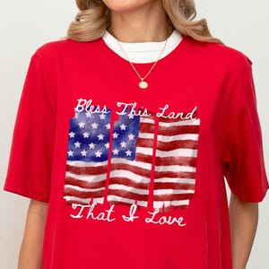 May include: Red t-shirt with a graphic of four American flags and the text "Bless this land that I love."