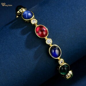 Op de afbeelding: Een gouden armband met een reeks kleurrijke edelstenen. De armband bevat een blauwe ster-saffier, een rode robijn, een groene smaragd en verschillende heldere, ronde diamanten. De armband is geplaatst tegen een donkerblauwe achtergrond.