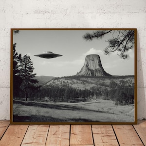 May include: Monochrome framed print depicting a UFO above Devil's Tower in Wyoming. The scene includes a grassy field, a forest, and a large rock formation under a cloudy sky. The print is displayed on a wooden floor.
