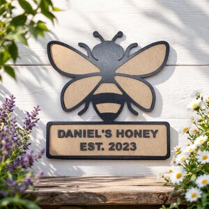 May include: A wooden bee-shaped sign with a black outline and tan-colored wings and body. Below the bee is a rectangular sign with the text "DANIEL'S HONEY EST. 2023" in black lettering. The sign is mounted on a white wooden surface.