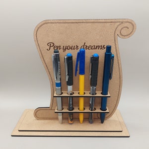 May include: A wooden pen holder with the phrase "Pen your dreams" engraved on it. The holder has a scroll design and holds five pens of various colors, including silver, black, yellow, and blue. The pens are displayed in a neat row.