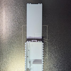 May include: A white, rectangular, open-top container with a textured, ridged border. The lid is separate and rectangular. The container is on a dark, textured surface.