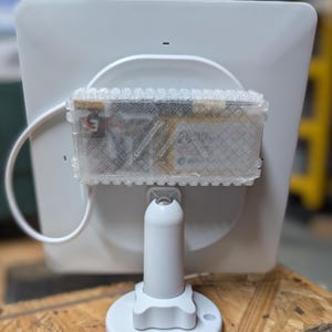 May include: A white, square-shaped electronic device with a clear, rectangular component attached to the back. The device is mounted on a white, adjustable stand with a circular base. The background is a wooden surface.