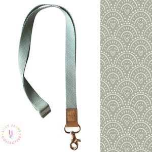 May include: A pale green lanyard with a gold-coloured clasp and a small leather tag. The tag is embossed with the text "Lilly Jayne Collective". The lanyard is made of a woven fabric. The background features a repeating pattern of white arches and hearts on a green background.