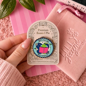 May include: A round enamel pin with a scalloped gold edge featuring a light blue background, a dark blue butterfly shape, and a bright pink open book. The text reads antisocial BUTTERFLY. It is mounted on a decorative cream-coloured card.