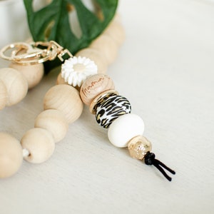 May include: A wooden beaded keychain with a gold key ring, a white flower charm, a wooden bead engraved with the word "Mama", a black and white leopard print bead, a white bead, and a gold bead with a black tassel.