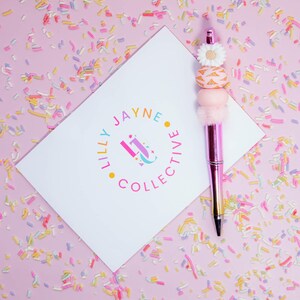 May include: A white card with the text "Lilly Jayne Collective" in a colourful circle logo. A pink pen with a white flower and pink and gold beads on the top is on the card. The card and pen are on a pink background with colourful sprinkles.