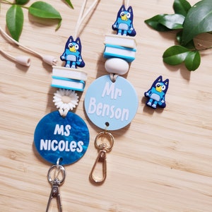 Pu&ograve; includere: Due lacci blu e bianchi con un personaggio di cane cartone animato blu, una pila di libri e un ciondolo a forma di fiore bianco. I lacci hanno un cerchio blu con testo bianco che recita "Ms. Nicoles" e "Mr. Benson".