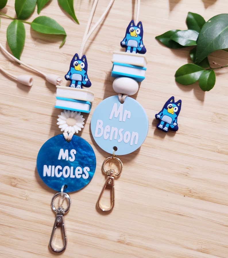 Pu&ograve; includere: Due lacci blu e bianchi con un personaggio di cane cartone animato blu, una pila di libri e un ciondolo a forma di fiore bianco. I lacci hanno un cerchio blu con testo bianco che recita "Ms. Nicoles" e "Mr. Benson".