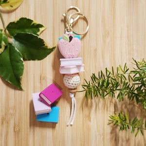May include: A pink apple-shaped keyring with the word "TEACH" on it. The keyring has a stack of three small books, a white crocheted ball, and a white tassel. The keyring is attached to a gold split ring.