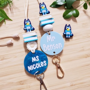 Pu&ograve; includere: Due lacci blu e bianchi con un personaggio di cane cartone animato blu, una pila di libri e un ciondolo di margherita bianca. I lacci hanno un cerchio blu con testo bianco che recita "Ms. Nicoles" e "Mr. Benson".