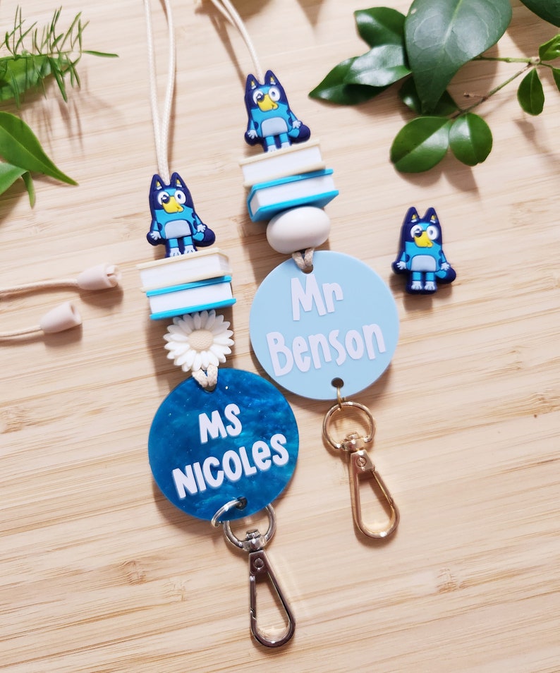 Pu&ograve; includere: Due lacci blu e bianchi con un personaggio di cane cartone animato blu, una pila di libri e un ciondolo di margherita bianca. I lacci hanno un cerchio blu con testo bianco che recita "Ms. Nicoles" e "Mr. Benson".