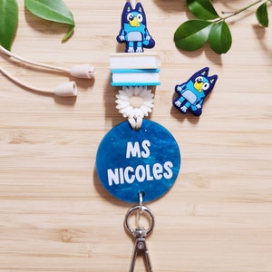 Pu&ograve; includere: Un laccio blu e bianco con un'etichetta rotonda blu che dice "MS NICOLES" e un ciondolo a forma di cane di cartone animato blu. Il laccio ha un moschettone argentato e un ciondolo a forma di margherita bianca.