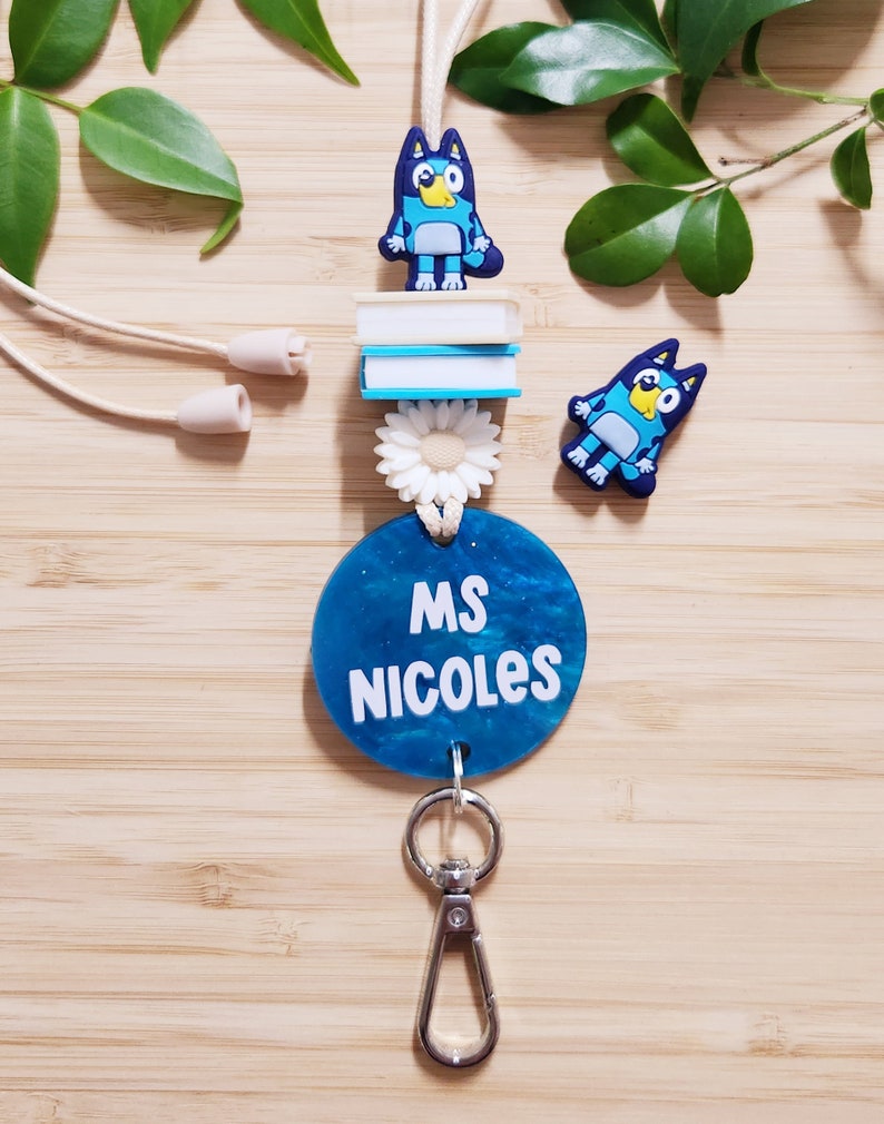 Pu&ograve; includere: Un laccio blu e bianco con un'etichetta rotonda blu che dice "MS NICOLES" e un ciondolo a forma di cane di cartone animato blu. Il laccio ha un moschettone argentato e un ciondolo a forma di margherita bianca.