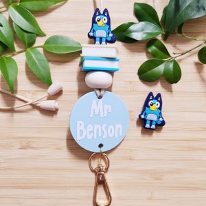 Pu&ograve; includere: Lacci blu e bianchi con un'etichetta rotonda blu che dice "Mr. Benson" e un ciondolo a forma di cane di cartone animato blu. Il laccio ha un moschettone dorato e una pila di libri blu e bianchi con un ciondolo a forma di cane di cartone animato blu in cima.