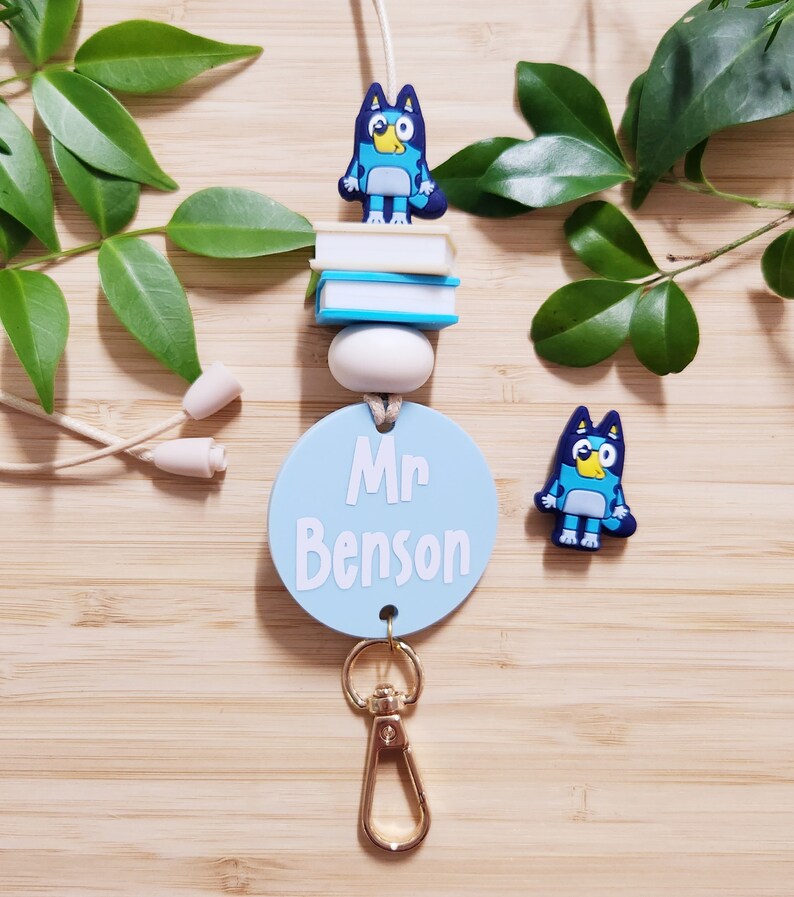 Pu&ograve; includere: Lacci blu e bianchi con un'etichetta rotonda blu che dice "Mr. Benson" e un ciondolo a forma di cane di cartone animato blu. Il laccio ha un moschettone dorato e una pila di libri blu e bianchi con un ciondolo a forma di cane di cartone animato blu in cima.