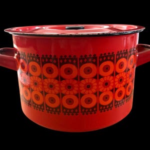Puede incluir: Una olla de cocina de esmalte rojo con tapa y dos asas laterales. La olla presenta un patrón repetitivo de diseños florales naranjas y marrones. El borde y las asas son de color marrón oscuro. Un artículo de cocina vintage.