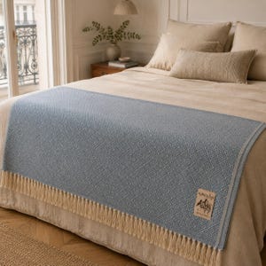 Puede incluir: Un camino de cama azul claro con un patrón de diamantes y flecos color crema. El camino de cama está colocado sobre una cama con ropa de cama y almohadas beige. Una etiqueta en el camino de cama dice "ALPACA Soft".