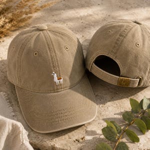 Puede incluir: Dos gorras de béisbol color caqui. Una gorra presenta un pequeño diseño de llama bordado en la parte delantera. La otra gorra está detrás, mostrando la correa ajustable con un logotipo de llama. Las gorras están hechas de una tela suave y texturizada.