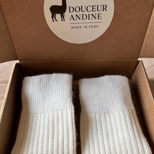 May include: A pair of off-white knitted socks inside a cardboard box. The socks have a ribbed design and a folded cuff. The box has a circular sticker with a llama silhouette and the text "DOUCEUR ANDINE MADE IN PERU."
