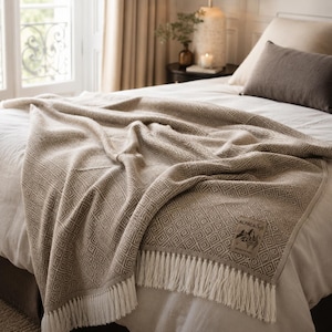 Könnte beinhalten: Eine beige Decke mit einem geometrischen Rautenmuster und Fransenbesatz. Die Decke ist über einem Bett drapiert und zeigt ihre Textur und ihr Design. Ein kleines Etikett mit Text und einer Berg-Grafik ist sichtbar.