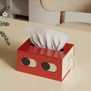 Peut inclure: Un distributeur de mouchoirs rouge avec une poignée blanche et deux motifs en forme d'yeux ovales. Le distributeur est rectangulaire et possède un intérieur blanc. Des mouchoirs blancs sortent du haut. Le tout est posé sur une surface claire.