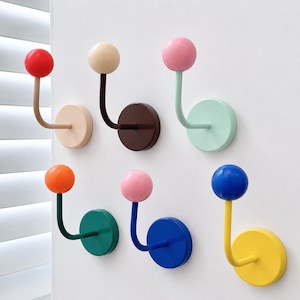Puede incluir: Un conjunto de seis ganchos de pared, cada uno con una base circular y un brazo curvo rematado con un pomo esférico. Los ganchos presentan una variedad de colores que incluyen rojo, beige, marrón, rosa, verde claro, naranja, verde oscuro, azul y amarillo.